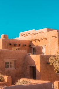 An adobe-style building features earthy tones and traditional vigas. The structure has a series of windows and a flat roof, creating a quintessential Southwestern architectural appearance. The building is illuminated by natural sunlight, casting distinct shadows and highlighting the rough texture of the walls. A bright, clear blue sky serves as the backdrop.