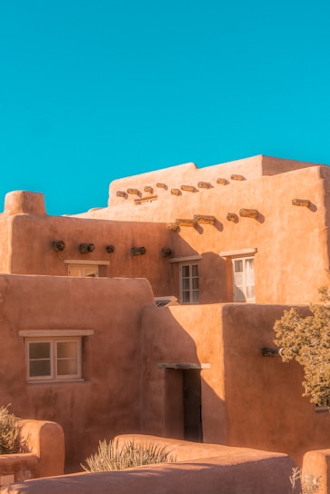 An adobe-style building features earthy tones and traditional vigas. The structure has a series of windows and a flat roof, creating a quintessential Southwestern architectural appearance. The building is illuminated by natural sunlight, casting distinct shadows and highlighting the rough texture of the walls. A bright, clear blue sky serves as the backdrop.