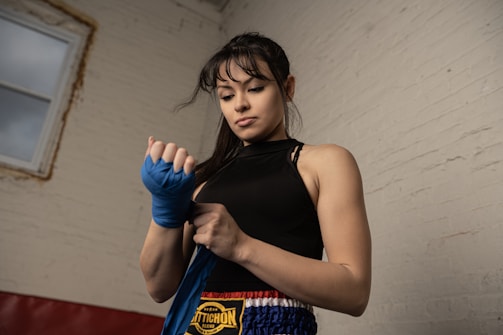 A woman in athletic clothing is wrapping her hands with blue boxing wraps in preparation for training. Her expression is focused and determined. The setting appears to be a gym with a rustic white brick wall and a window on the left side.