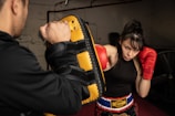 A woman wearing red boxing gloves is practicing punches with a coach holding a yellow and black focus mitt indoors. She appears focused and determined. The setting includes a gym-like environment with a dim background.