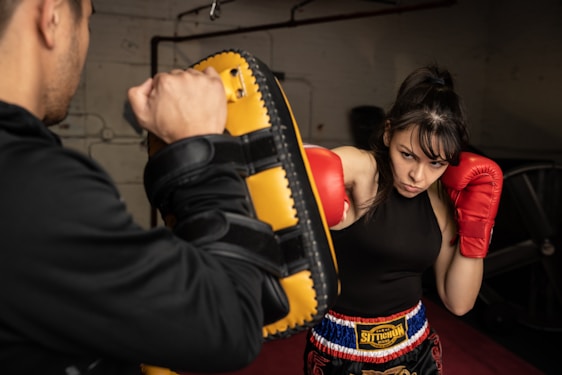 A woman wearing red boxing gloves is practicing punches with a coach holding a yellow and black focus mitt indoors. She appears focused and determined. The setting includes a gym-like environment with a dim background.