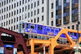 A bright blue and yellow train travels on an elevated track with background of modern city buildings. The train is branded with 'getir' and advertisements for groceries. The structure supporting the track is painted in shades of brown and orange.