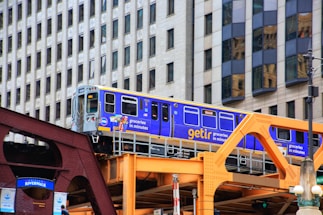 A bright blue and yellow train travels on an elevated track with background of modern city buildings. The train is branded with 'getir' and advertisements for groceries. The structure supporting the track is painted in shades of brown and orange.
