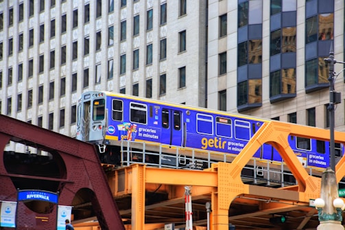 A bright blue and yellow train travels on an elevated track with background of modern city buildings. The train is branded with 'getir' and advertisements for groceries. The structure supporting the track is painted in shades of brown and orange.