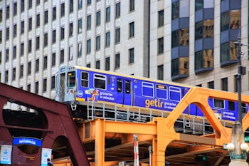A bright blue and yellow train travels on an elevated track with background of modern city buildings. The train is branded with 'getir' and advertisements for groceries. The structure supporting the track is painted in shades of brown and orange.