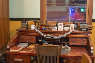 A vintage wooden desk adorned with various items, including books, a mug, greeting cards, and small decorations like baubles and a tinsel garland. The desk features multiple drawers and compartments and is situated against a window with the glow of indoor lights. An old-fashioned typewriter and a rotary calculator are also present, adding to the nostalgic ambiance.
