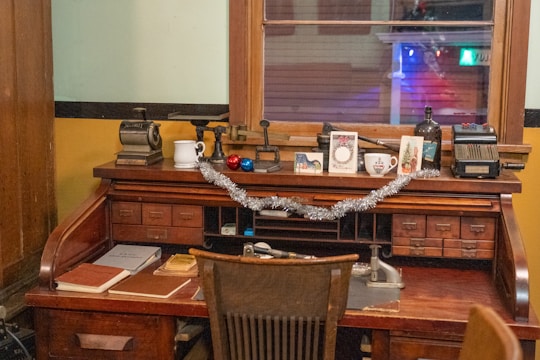 A vintage wooden desk adorned with various items, including books, a mug, greeting cards, and small decorations like baubles and a tinsel garland. The desk features multiple drawers and compartments and is situated against a window with the glow of indoor lights. An old-fashioned typewriter and a rotary calculator are also present, adding to the nostalgic ambiance.