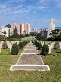 A cemetery with rows of gravestones aligned neatly along a pathway, set against a backdrop of urban high-rise buildings and a mountainous landscape. The sky is clear with few clouds.