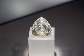 A sparkling diamond with a triangular cut is displayed on a simple white pedestal. The gem reflects a spectrum of colors against a neutral background.