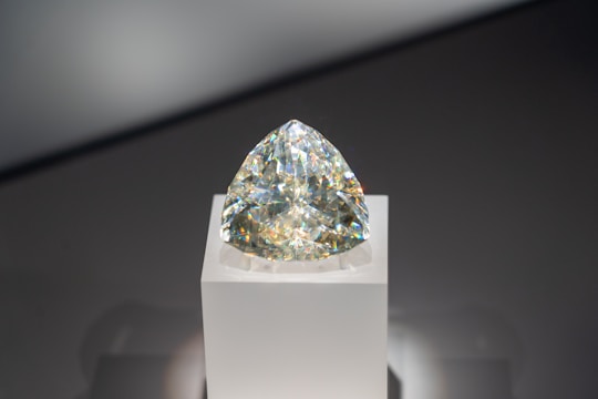 A sparkling diamond with a triangular cut is displayed on a simple white pedestal. The gem reflects a spectrum of colors against a neutral background.