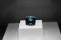 A dark blue rectangular gemstone is placed on a raised white platform against a dark background. The gem is faceted and exhibits a deep, rich hue, with light reflecting off its polished surfaces.