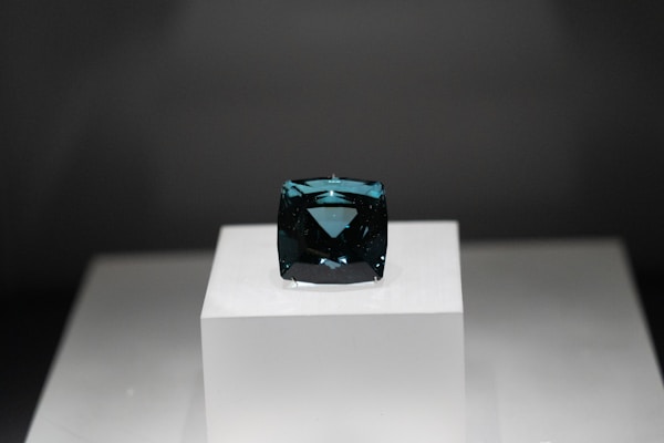 A dark blue rectangular gemstone is placed on a raised white platform against a dark background. The gem is faceted and exhibits a deep, rich hue, with light reflecting off its polished surfaces.