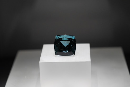 A dark blue rectangular gemstone is placed on a raised white platform against a dark background. The gem is faceted and exhibits a deep, rich hue, with light reflecting off its polished surfaces.