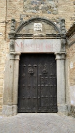 A stone archway with detailed carvings frames a large, ornate wooden door. The door features intricate metalwork and is surrounded by a rustic brick facade, highlighted by rocks and stones embedded in the upper section. An inscription in faded red text is visible above the arch.