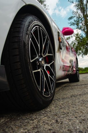 A sleek car is parked on a textured road surface, displaying a close-up view of its tire and alloy wheel. The car body features a unique design with graffiti-like artwork and vibrant colors, including a noticeable pink side mirror. In the background, tall trees reach up to the cloudy sky, adding a natural element to the scene.