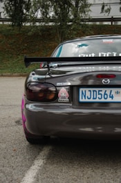 The rear view of a dark gray sports car with a prominent spoiler. Decorative stickers are visible on the back, including a character and the phrase 'DRIVE SAFE'. The car has round tail lights and is parked on an asphalt road next to some greenery.
