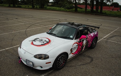 A customized sports car is parked in an empty parking lot. The car features vibrant anime-style graphics predominantly in pink and white, with some black elements, including Japanese characters on the hood and sides. The car is a convertible with a visible rear spoiler and black wheels displaying a design element in red.