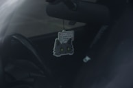 A car interior with a cute cat-themed air freshener hanging from the rearview mirror. The air freshener has a cartoon cat design with text on it.