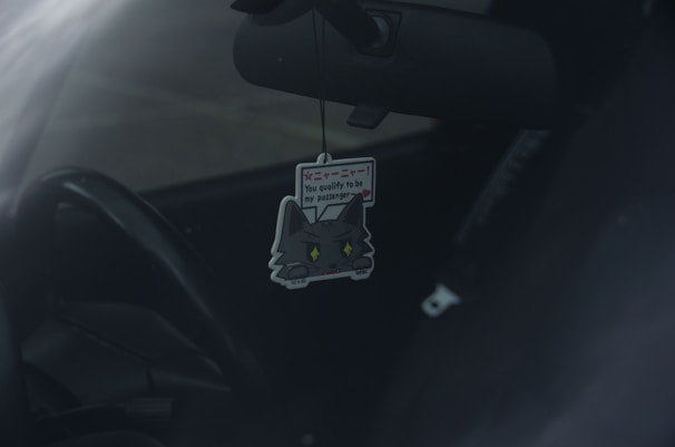 A car interior with a cute cat-themed air freshener hanging from the rearview mirror. The air freshener has a cartoon cat design with text on it.