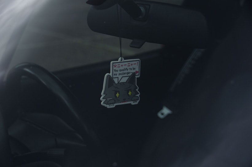 A car interior with a cute cat-themed air freshener hanging from the rearview mirror. The air freshener has a cartoon cat design with text on it.