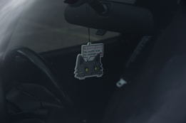 A car interior with a cute cat-themed air freshener hanging from the rearview mirror. The air freshener has a cartoon cat design with text on it.