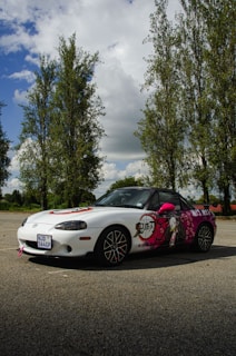 A white sports car with artistic decals featuring anime-style graphics is parked on an outdoor surface surrounded by tall trees under a partly cloudy sky.