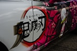 A car features an anime-themed wrap with vibrant pink and white colors. The artwork includes stylized Japanese kanji characters and a depiction of a female anime character with long hair, a bamboo gag, and wearing a kimono. Pink cherry blossom flowers are scattered around the design, adding a decorative touch.