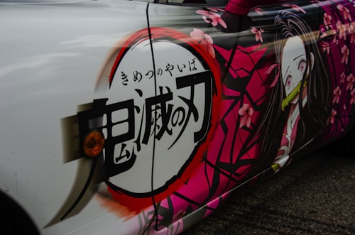 A car features an anime-themed wrap with vibrant pink and white colors. The artwork includes stylized Japanese kanji characters and a depiction of a female anime character with long hair, a bamboo gag, and wearing a kimono. Pink cherry blossom flowers are scattered around the design, adding a decorative touch.
