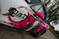 A car with an anime-themed design featuring Japanese characters and an animated character on the side. The design is vibrant, with cherry blossom motifs and a predominantly pink and white color scheme. The car is parked on a road, with trees and a clear sky in the background.