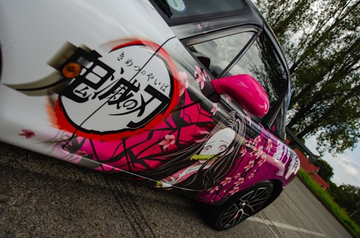 A car with an anime-themed design featuring Japanese characters and an animated character on the side. The design is vibrant, with cherry blossom motifs and a predominantly pink and white color scheme. The car is parked on a road, with trees and a clear sky in the background.
