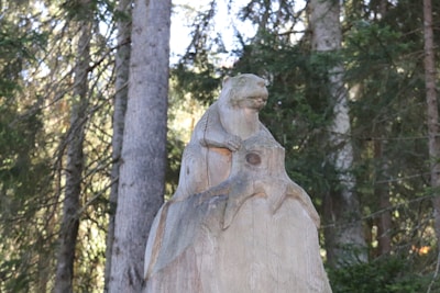 A wooden sculpture of an animal resembling a beaver or marmot is perched on a carved tree stump. The setting is a dense forest with tall trees and thick foliage in the background, suggesting a natural and rustic environment.