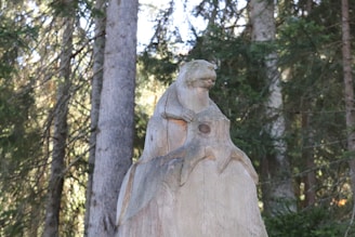 A wooden sculpture of an animal resembling a beaver or marmot is perched on a carved tree stump. The setting is a dense forest with tall trees and thick foliage in the background, suggesting a natural and rustic environment.