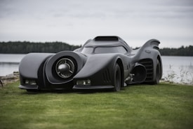 A sleek, futuristic black car with large distinctive fins and an aerodynamic design is parked on a grassy area near a body of water. It features a prominent central turbine on the front, evoking a sense of power and innovation. In the background, there is a calm lake and a dense forest under a cloudy sky, creating a serene yet mysterious atmosphere.