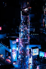 A vibrant urban scene featuring a bustling city street at night, illuminated by bright neon lights and advertisements. The street is filled with numerous cars and people, and tall buildings adorned with billboards line both sides.