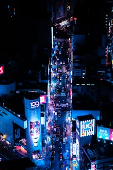 A vibrant urban scene featuring a bustling city street at night, illuminated by bright neon lights and advertisements. The street is filled with numerous cars and people, and tall buildings adorned with billboards line both sides.