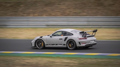 A sleek, high-performance sports car speeds along a race track with precision and agility. The car has a light gray body with distinctive black accents and a large rear wing. The background features a blurred raceway and safety barriers, emphasizing the car's speed.
