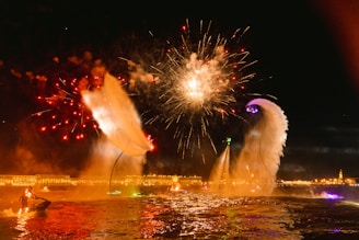 Fireworks light up the night sky above a brightly lit body of water. People are performing water stunts using jet-packs or water-powered hoverboards, creating large plumes of water. Buildings with illuminated facades are visible in the background, adding to the festive atmosphere.