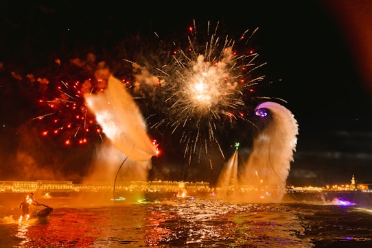 Fireworks light up the night sky above a brightly lit body of water. People are performing water stunts using jet-packs or water-powered hoverboards, creating large plumes of water. Buildings with illuminated facades are visible in the background, adding to the festive atmosphere.