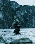 A person wearing a green raincoat and backpack stands in shallow, clear water, holding a fishing rod. The background features steep, rocky cliffs covered with dense greenery, creating a serene and rugged environment.
