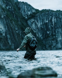 A person wearing a green raincoat and backpack stands in shallow, clear water, holding a fishing rod. The background features steep, rocky cliffs covered with dense greenery, creating a serene and rugged environment.