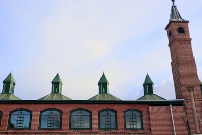 A brick building with an architectural design featuring arched windows and a series of conical green roofs. A tall brick tower extends from the right side of the building, topped with a small steeple.
