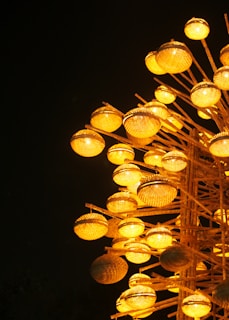 A cluster of illuminated wicker baskets arranged on wooden poles, casting a warm yellow glow against a dark background. The spherical baskets are intricately woven and emit light, creating an artistic display.