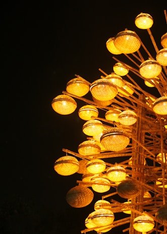 A cluster of illuminated wicker baskets arranged on wooden poles, casting a warm yellow glow against a dark background. The spherical baskets are intricately woven and emit light, creating an artistic display.