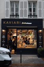 A delicatessen storefront with a large glass window displays a variety of food items. Inside, several people are seen shopping or working. The exterior features a dark facade with gold and white lettering, and the warm lighting from inside creates a cozy atmosphere.