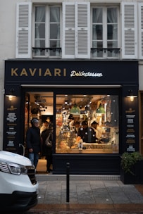 A delicatessen storefront with a large glass window displays a variety of food items. Inside, several people are seen shopping or working. The exterior features a dark facade with gold and white lettering, and the warm lighting from inside creates a cozy atmosphere.