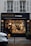 A delicatessen storefront with a large glass window displays a variety of food items. Inside, several people are seen shopping or working. The exterior features a dark facade with gold and white lettering, and the warm lighting from inside creates a cozy atmosphere.