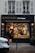 A delicatessen storefront with a large glass window displays a variety of food items. Inside, several people are seen shopping or working. The exterior features a dark facade with gold and white lettering, and the warm lighting from inside creates a cozy atmosphere.