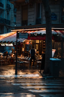 A cozy outdoor café scene with several people seated at tables under a red and white striped awning. The ambience is enhanced by warm lighting reflecting on wet pavement, suggesting recent rain. A street sign indicates the name Avenue de Bourdonnais, and decorations with greenery create a festive atmosphere.