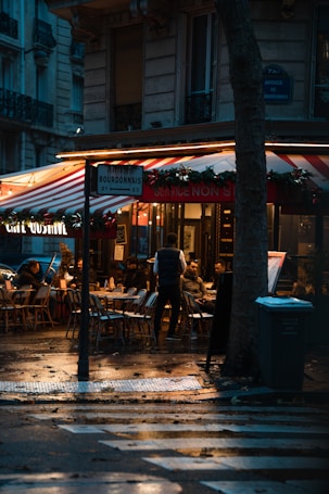 A cozy outdoor caf&eacute; scene with several people seated at tables under a red and white striped awning. The ambience is enhanced by warm lighting reflecting on wet pavement, suggesting recent rain. A street sign indicates the name Avenue de Bourdonnais, and decorations with greenery create a festive atmosphere.