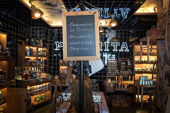 A cozy and rustic store interior with wooden shelves filled with various bottles and jars on display. A chalkboard sign stands in the center, listing items like Ibérico charcuterie and wines. Warm lighting enhances the inviting atmosphere, and a black tiled wall features illuminated text in the background.